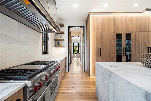 kitchen featuring wall chimney range hood, open shelves, light stone counters, light wood-style flooring, and range with two ovens