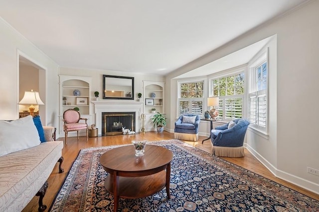 living room featuring built in shelves, baseboards, a fireplace with flush hearth, and wood finished floors