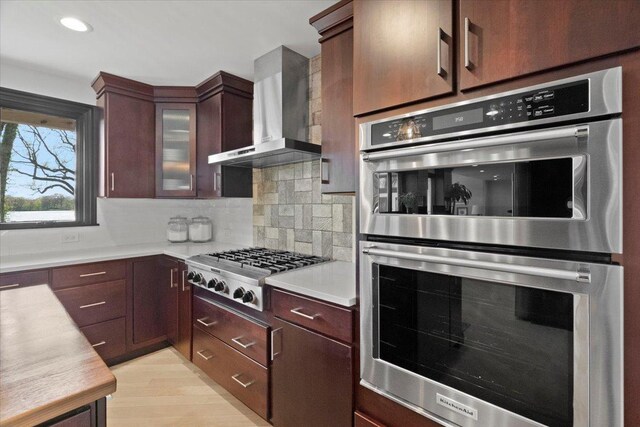 kitchen featuring wall chimney range hood, decorative backsplash, appliances with stainless steel finishes, light countertops, and glass insert cabinets