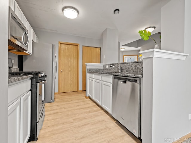 kitchen featuring appliances with stainless steel finishes, a sink, light wood finished floors, dark stone counters, and white cabinetry