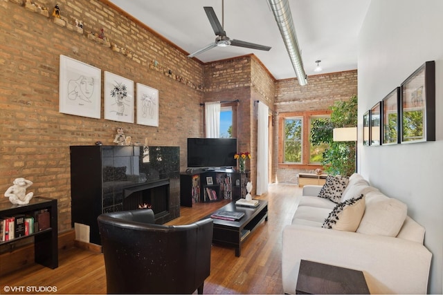 living area with a warm lit fireplace, wood finished floors, a ceiling fan, brick wall, and a high ceiling