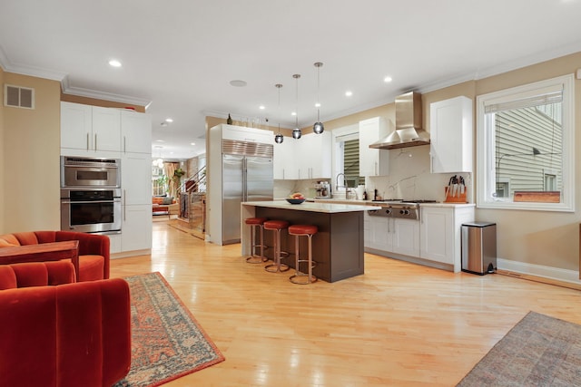 kitchen featuring wall chimney exhaust hood, a breakfast bar, stainless steel appliances, light wood-style floors, and ornamental molding