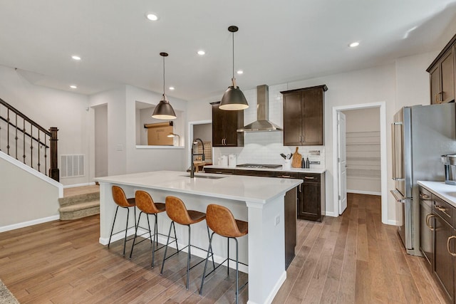 kitchen featuring wall chimney range hood, dark brown cabinets, a sink, stainless steel appliances, and light wood-type flooring