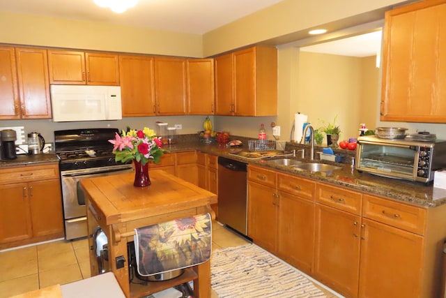 kitchen with stainless steel appliances, a sink, light tile patterned floors, brown cabinets, and dark stone counters