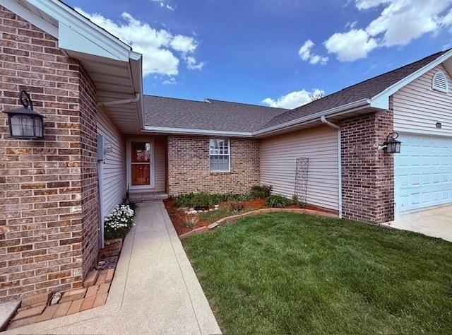 view of exterior entry with brick siding, an attached garage, roof with shingles, and a yard