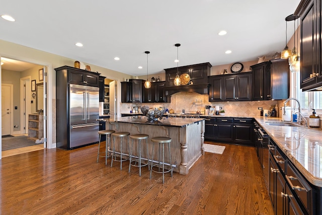 kitchen featuring a kitchen island, built in fridge, a sink, dark wood-style floors, and dark stone counters