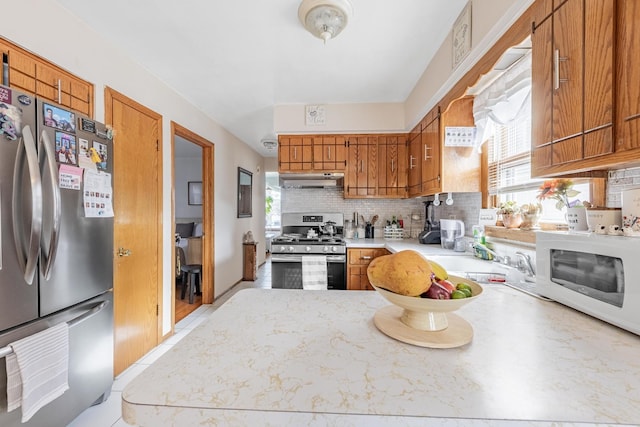 kitchen featuring brown cabinetry, decorative backsplash, under cabinet range hood, a sink, and stainless steel appliances