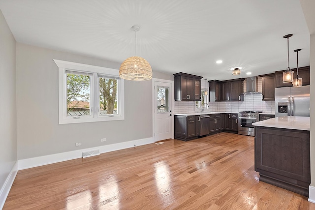 kitchen featuring dark brown cabinetry, baseboards, wall chimney range hood, light countertops, and stainless steel appliances