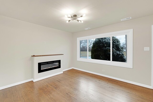 unfurnished living room with visible vents, wood finished floors, baseboards, a chandelier, and a glass covered fireplace
