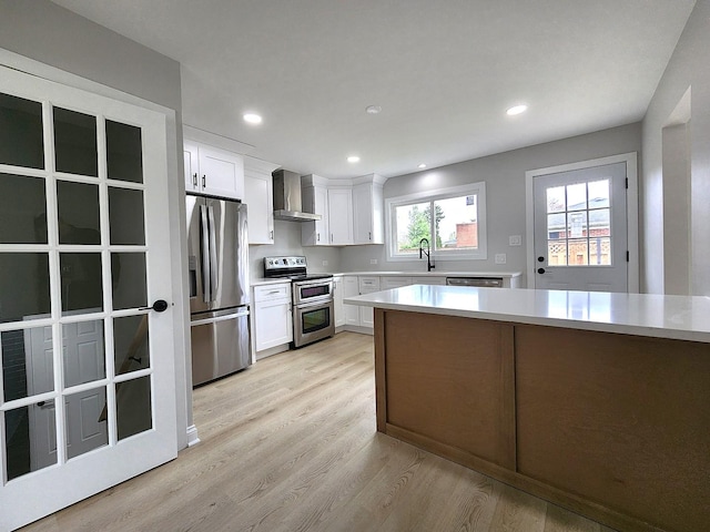 kitchen featuring wall chimney range hood, light countertops, a sink, light wood finished floors, and appliances with stainless steel finishes