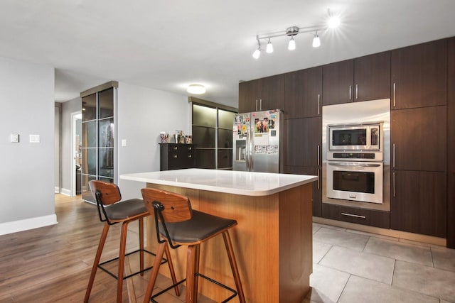 kitchen featuring dark brown cabinets, appliances with stainless steel finishes, a breakfast bar, light countertops, and a kitchen island