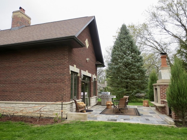 view of side of property featuring a patio, roof with shingles, a chimney, brick siding, and an outdoor brick fireplace