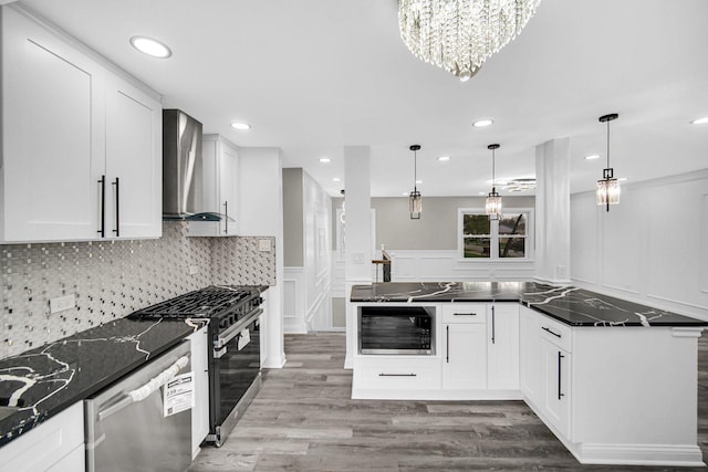 kitchen featuring wall chimney range hood, appliances with stainless steel finishes, a decorative wall, a chandelier, and wood finished floors