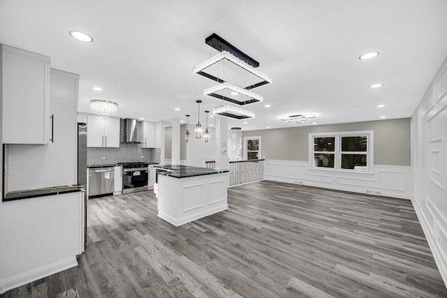 kitchen with wall chimney exhaust hood, stainless steel appliances, dark countertops, a chandelier, and white cabinets
