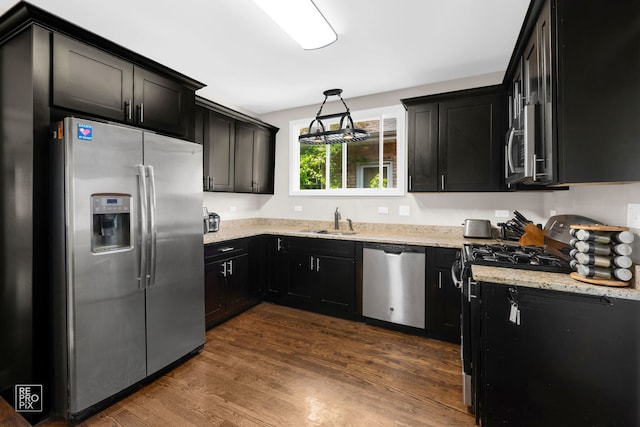 kitchen with appliances with stainless steel finishes, a sink, dark cabinetry, light stone counters, and dark wood-type flooring