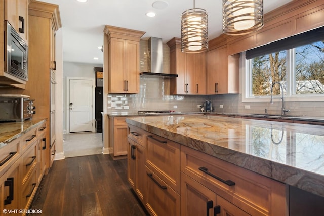 kitchen featuring wall chimney range hood, light stone counters, a sink, stainless steel appliances, and tasteful backsplash