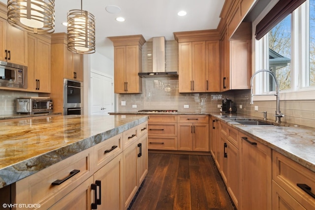 kitchen with light stone counters, appliances with stainless steel finishes, a sink, wall chimney exhaust hood, and tasteful backsplash