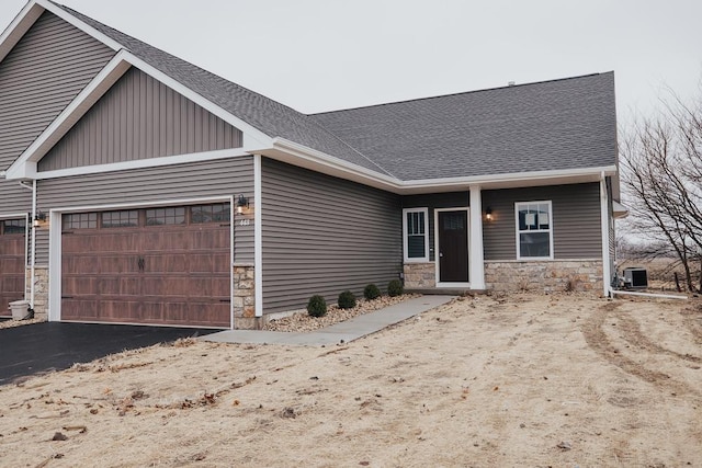 view of front of property featuring an attached garage, a shingled roof, central air condition unit, and stone siding