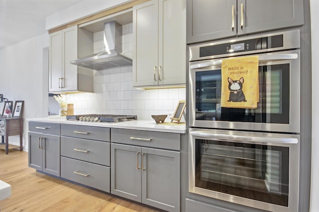kitchen featuring light wood-style floors, wall chimney range hood, backsplash, gray cabinetry, and stainless steel appliances