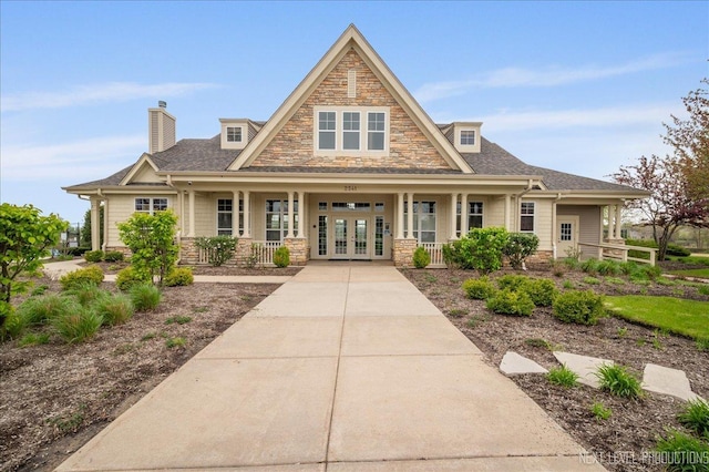 view of front of property featuring french doors, covered porch, stone siding, a chimney, and roof with shingles