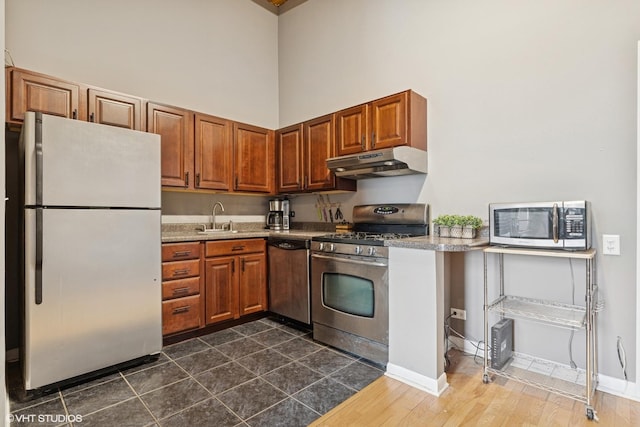kitchen featuring under cabinet range hood, a sink, brown cabinets, appliances with stainless steel finishes, and a high ceiling
