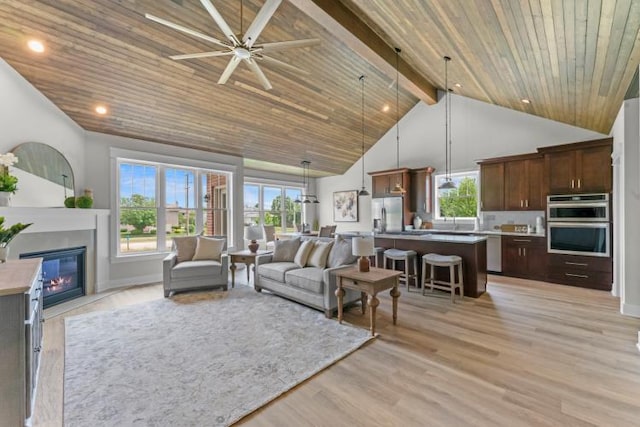 living room with a glass covered fireplace, beamed ceiling, ceiling fan, wood ceiling, and light wood finished floors