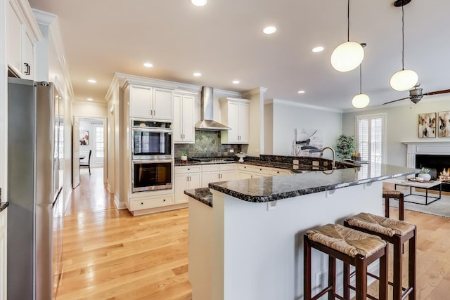 kitchen featuring appliances with stainless steel finishes, crown molding, wall chimney exhaust hood, a breakfast bar area, and light wood-style flooring