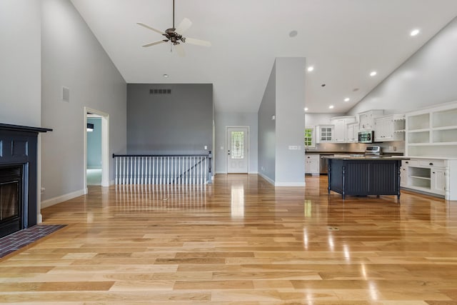 unfurnished living room featuring high vaulted ceiling, a ceiling fan, a fireplace with flush hearth, light wood-style floors, and baseboards