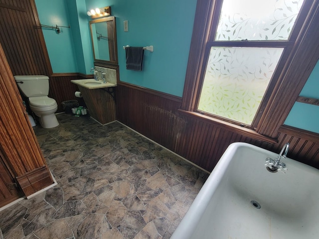bathroom with wood walls, toilet, a wainscoted wall, stone finish floor, and a freestanding bath