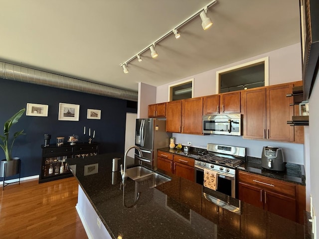 kitchen with a sink, brown cabinetry, wood finished floors, rail lighting, and appliances with stainless steel finishes