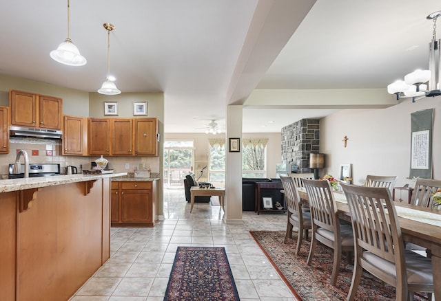 kitchen featuring under cabinet range hood, brown cabinetry, light stone counters, ceiling fan with notable chandelier, and decorative backsplash