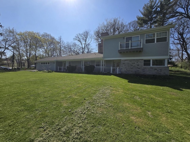 rear view of property featuring a lawn, stone siding, a chimney, and a balcony
