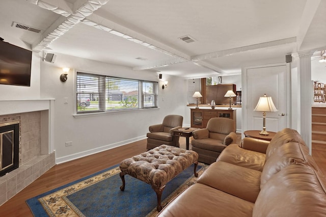 living area featuring a tiled fireplace, baseboards, wood finished floors, beamed ceiling, and ornate columns