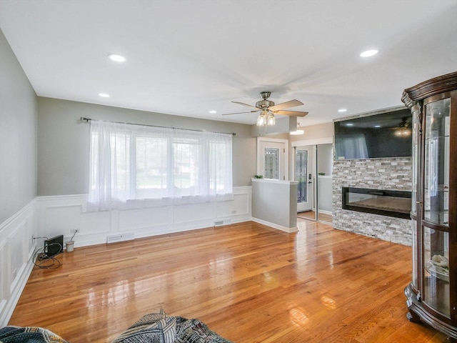 unfurnished living room featuring wood finished floors, visible vents, a ceiling fan, wainscoting, and a stone fireplace
