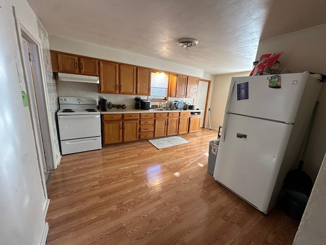 kitchen featuring white appliances, brown cabinetry, light wood finished floors, under cabinet range hood, and a sink
