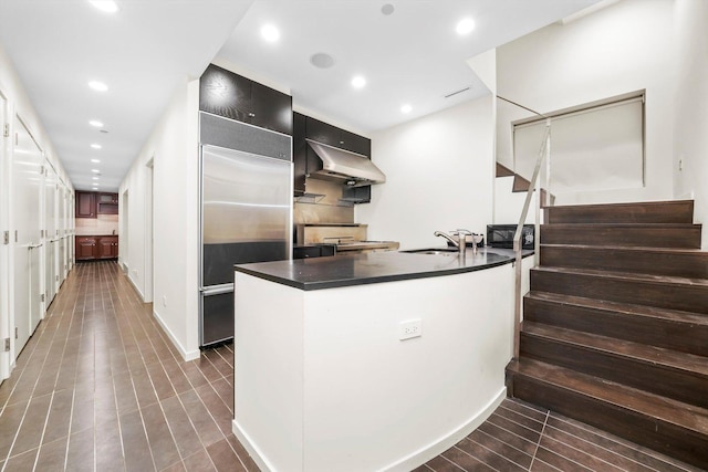 kitchen with under cabinet range hood, dark countertops, built in refrigerator, and a sink