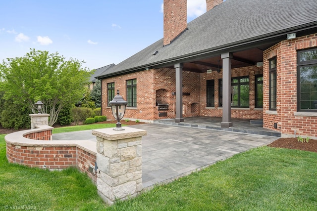 exterior space with a shingled roof, brick siding, a chimney, and a patio