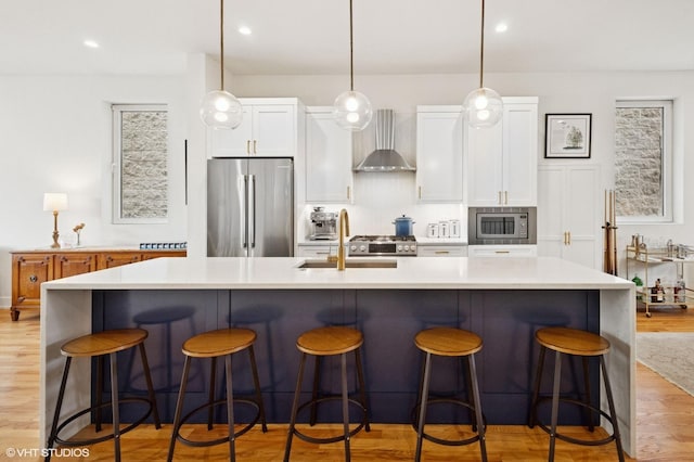 kitchen featuring stainless steel appliances, wall chimney range hood, a sink, light wood-type flooring, and white cabinets