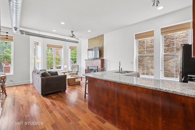 kitchen featuring a ceiling fan, wood-type flooring, a sink, a glass covered fireplace, and light stone countertops