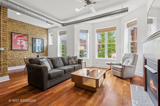living area featuring brick wall, wood-type flooring, baseboards, a ceiling fan, and a lit fireplace