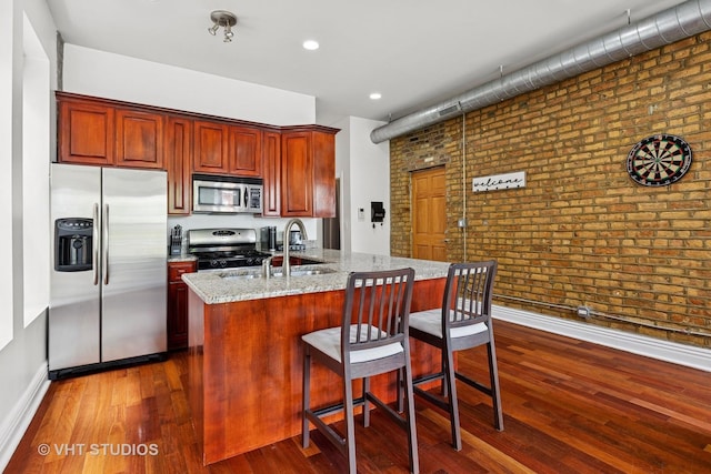 kitchen with appliances with stainless steel finishes, brick wall, baseboards, dark wood-style flooring, and a kitchen island with sink