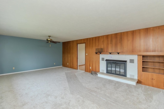 unfurnished living room with ceiling fan, a brick fireplace, carpet flooring, wooden walls, and baseboards