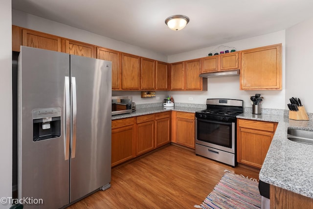 kitchen featuring stainless steel appliances, under cabinet range hood, light wood finished floors, a sink, and brown cabinetry