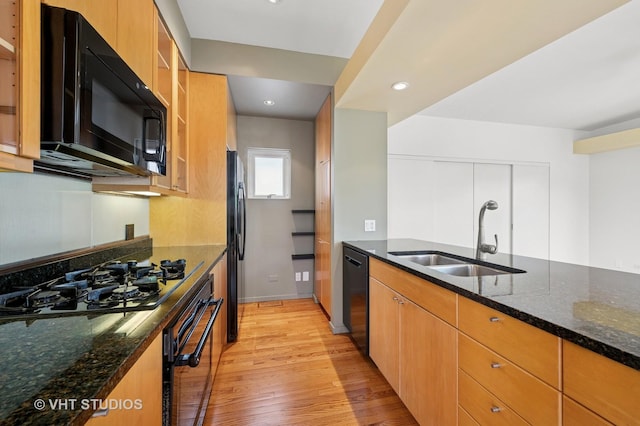 kitchen featuring black appliances, a sink, light wood finished floors, dark stone counters, and glass insert cabinets