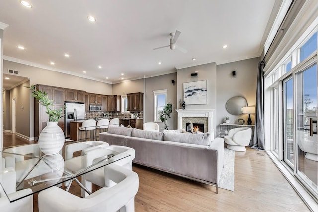 living room featuring a ceiling fan, crown molding, a warm lit fireplace, light wood finished floors, and recessed lighting