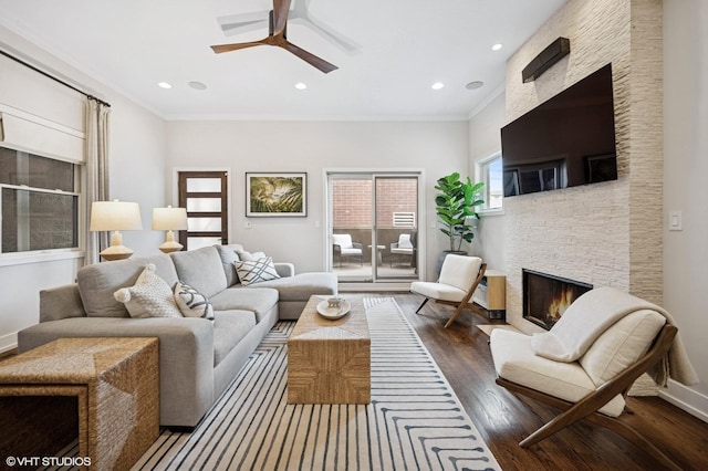 living room featuring a stone fireplace, wood finished floors, a ceiling fan, crown molding, and recessed lighting