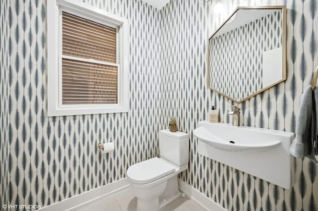 half bathroom with wallpapered walls, toilet, tile patterned floors, baseboards, and a sink