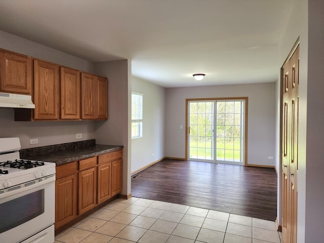 kitchen featuring white gas range, under cabinet range hood, light tile patterned flooring, brown cabinets, and dark countertops