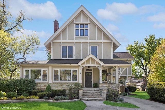 tudor house featuring a chimney, stucco siding, covered porch, stone siding, and a front lawn
