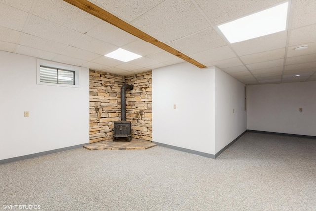 basement featuring a wood stove, baseboards, a drop ceiling, and carpet